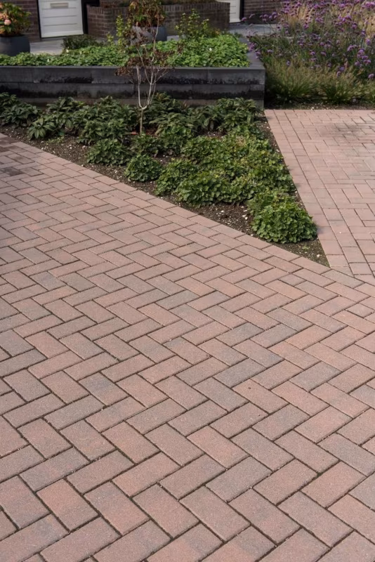 Oprit van roodbruine klinkers in visgraatverband langs plantvak met struiken en zwarte randafwerking