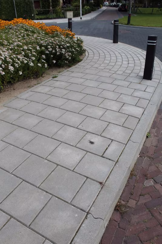 Gebogen voetpad van grijze betontegels langs bloemenborder en zwarte paaltjes