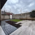 Achtertuin met visgraatpad en lichtgrijze tegels langs gazon en overkapping, nat na regen