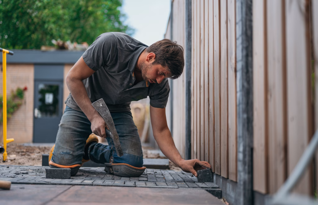 Hovenier legt sierbestrating aan in een zonnige tuin, met gebruik van diverse soorten stenen en patronen, ter illustratie van de kwaliteit en veelzijdigheid van producten aangeboden door Bestratingsmarkt.com.