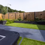 Gazon met zwarte trampolinehoes en GeoCeramica Flow Black tegels langs houten schutting