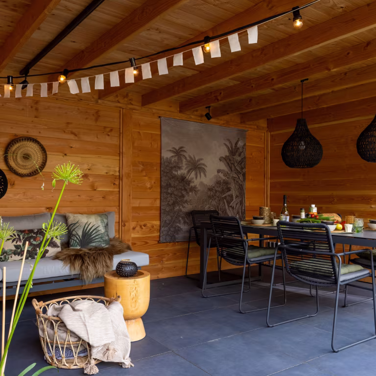 Gedekte eettafel met zwarte stoelen en zitbank onder houten overkapping met slingers