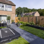 Achtertuin met GeoCeramica Flow Black tegels, ingegraven trampoline, gazon en bakstenen huis