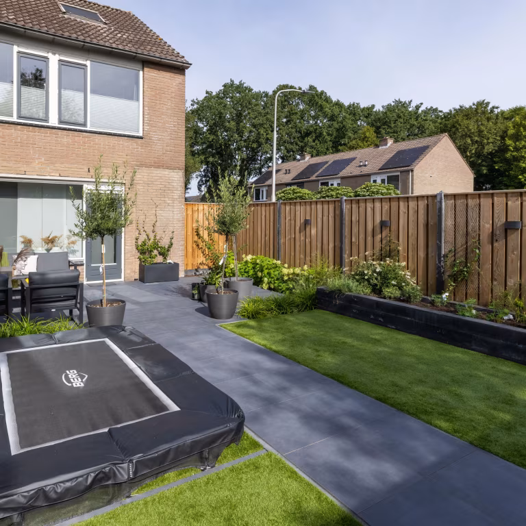 Achtertuin met GeoCeramica Flow Black tegels, ingegraven trampoline, gazon en bakstenen huis