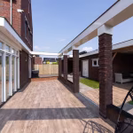 Lange doorloop met Timber Noce houtlooktegels tussen woning en bakstenen pilaren, zonnige dag