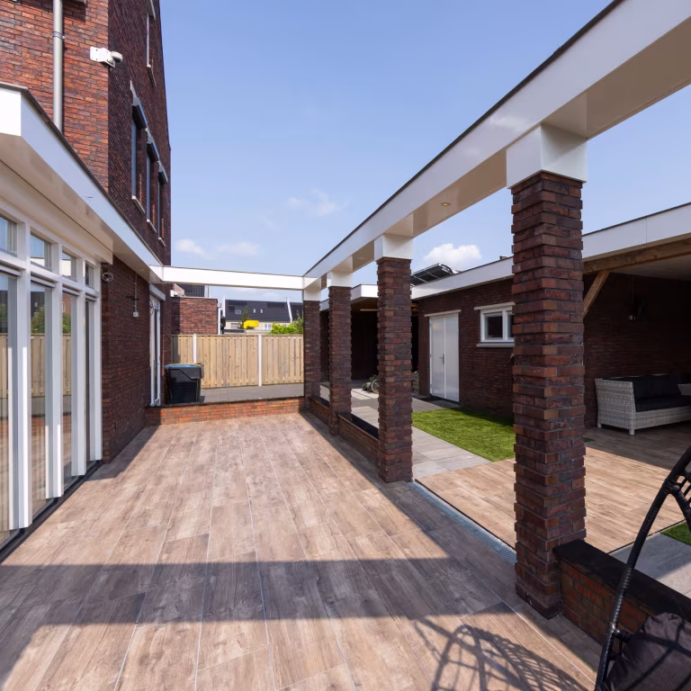 Lange doorloop met Timber Noce houtlooktegels tussen woning en bakstenen pilaren, zonnige dag