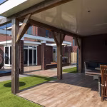 Onder overkapping uitzicht op Timber Noce terras, kunstgras en bakstenen woning met pui