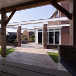 Doorkijk vanuit houten overkapping over terras met Timber Noce tegels en bakstenen pilaren