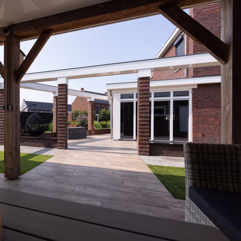 Doorkijk vanuit houten overkapping over terras met Timber Noce tegels en bakstenen pilaren
