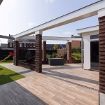 Terras van Timber Noce tegels met witte dakrand, bakstenen pilaren en speeltoestel op achtergrond