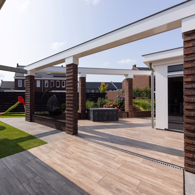 Terras van Timber Noce tegels met witte dakrand, bakstenen pilaren en speeltoestel op achtergrond