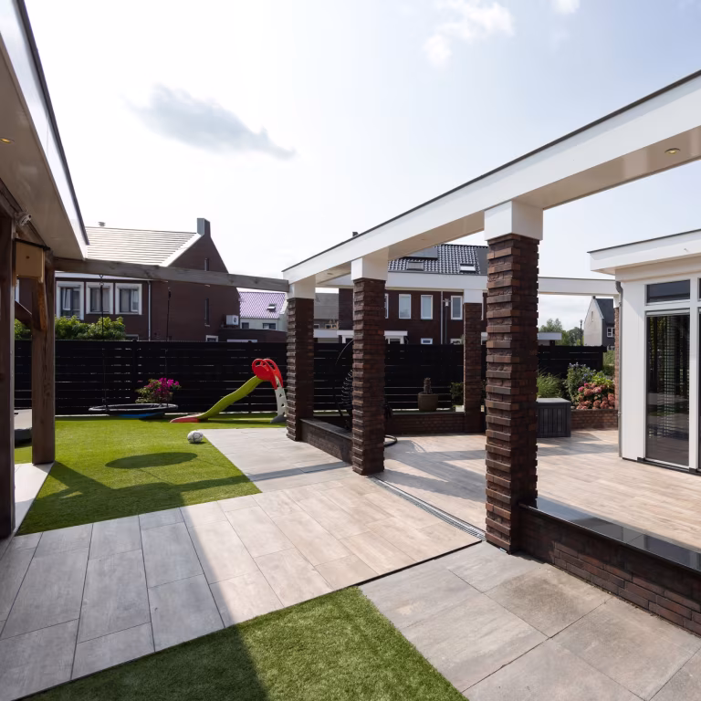 Terras met Timber Noce tegels, kunstgras, speeltoestel en bakstenen pilaren onder witte dakrand