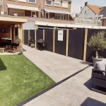 Achtertuin met Kera Full Body Nice tegels, overkapping, kunstgras en zwarte schutting