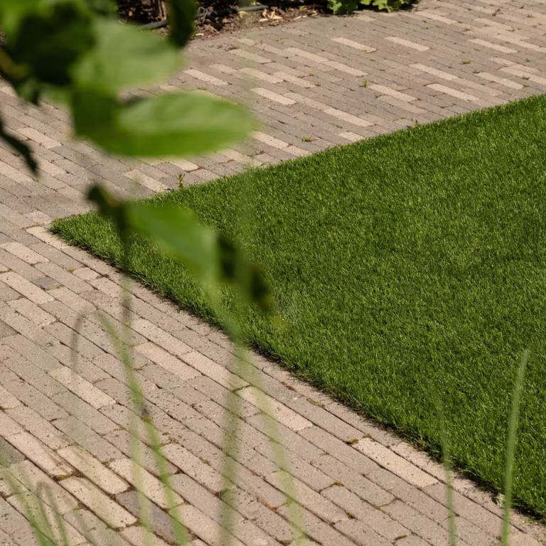Hoek van kunstgras omlijst door lichte waalformaat klinkers met groen blad op voorgrond