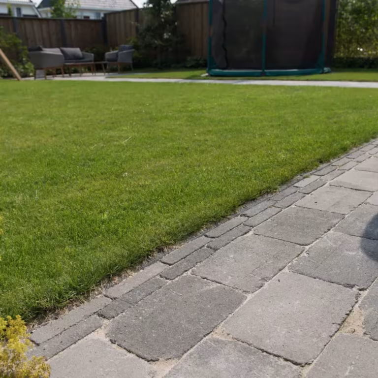 Tambour Smook stenenpad langs gazon met tuinmeubels en trampoline op achtergrond