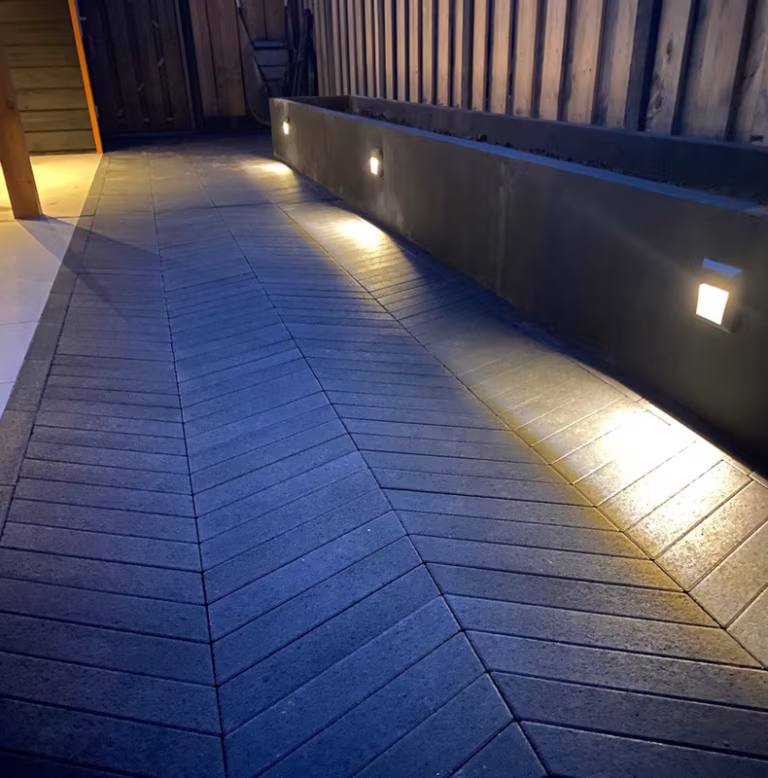 Visgraatpad Romano Punto Nero langs betonnen muur met inbouwverlichting in de avond