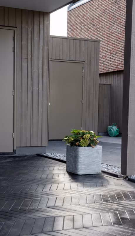 Zwarte visgraatbestrating Romano Punto Nero bij grijze berging met plantenbak en bloemen
