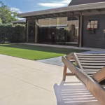 Terras met Ceramaxx Andes Gold tegels, houten ligstoel en tuinhuis met gras