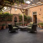 Binnenplaats met Cerasun Siena tegels, pergola, rieten fauteuils en vuurtafel bij bakstenen muur