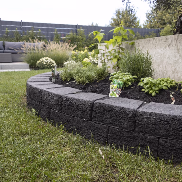 Ronde plantenbak van Pilestone Coal langs gazon met siergrassen en hosta's
